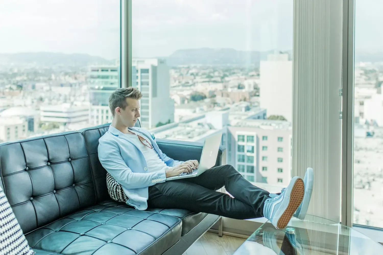 Mann sitzt mit Laptop auf einem Sofa vor großen Fenstern mit Blick über die Stadt